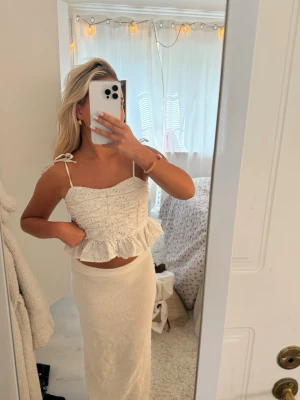 Vit broderad linnetopp med volang - Söt vit linnetopp med broderade detaljer och volangkant nedtill. Toppen har smala axelband som knyts på axlarna och en croppad passform. Perfekt för sommaren och ger en romantisk vibe.