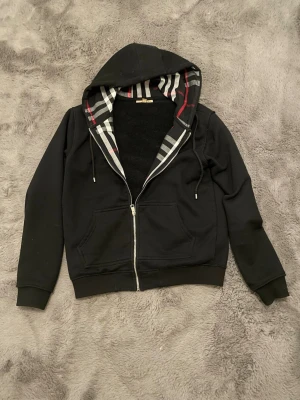 Svart zip hoodie från Burberry - Snygg svart hoodie från Burberry med dragkedja och klassiskt rutigt foder i huvan. Två fickor framtill och justerbar dragsko i huvan. Perfekt för dig som gillar stilrena plagg med exklusiv känsla. Materialet känns mjukt och bekvämt.