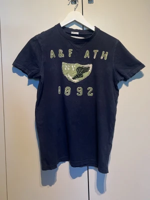 Mörkblå t-shirt Abercrombie & Fitch - Säljer en mörkblå t-shirt från Abercrombie & Fitch med broderade detaljer och text på bröstet. T-shirten har korta ärmar och ett coolt patch-märke med vinge och siffror. Perfekt för dig som gillar klassisk college-stil.