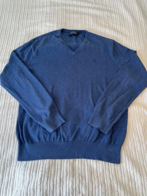  Polo Ralph Lauren tröja - Snygg blå stickad tröja från Polo Ralph Lauren med V-ringning och diskret logga på bröstet. Tröjan har långa ärmar och ribbstickade muddar vid ärmslut och nederkant. Perfekt för att hålla värmen under kyliga dagar.