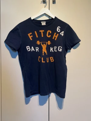 Marinblå t-shirt Abercrombie & Fitch - Marinblå t-shirt från Abercrombie & Fitch med tryck i vitt och orange framtill. Motiv med texten 'Fitch Bar Keg Club' och en figur som lyfter skivstång. Klassisk rund hals och korta ärmar. Tillverkad i mjuk bomull.