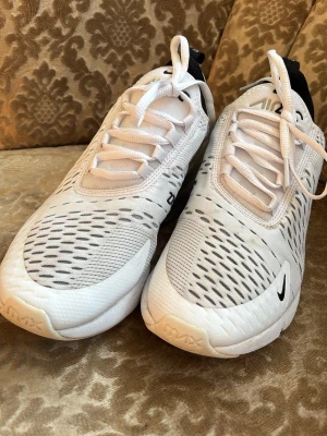 Nike Air Max 270 vita sneakers - Säljer ett par Nike Air Max 270 sneakers i vitt med svarta detaljer och mesh-paneler för extra ventilation. Skorna har en chunky sula med Air Max-dämpning och klassisk Nike-logga på sidan.