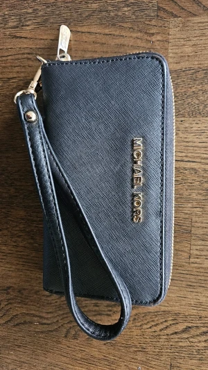 Svart plånbok från Michael Kors - Stilren svart plånbok från Michael Kors i strukturerat läder med guldfärgad logotyp och dragkedja. Plånboken har ett praktiskt handledsband, handledsbandet är inte orginal MK till denna plånbok. I använt men fortfarande fint skick, se bilder.