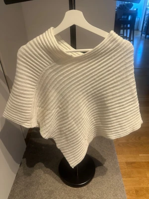 Vit ribbstickad poncho - Säljer en snygg vit poncho i ribbstickat material. Ponchon har en asymmetrisk form, perfekt att slänga över en tröja för extra värme. Enkel och stilren design som passar till många outfits.