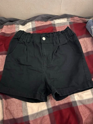 Svarta shorts med resårmidja - Säljer ett par svarta shorts med resår i midjan och knappstängning framtill. Shortsen har två fickor fram och två bakfickor. De är tillverkade i ett mjukt bomullsliknande material och har en avslappnad passform som passar perfekt till sommaren.