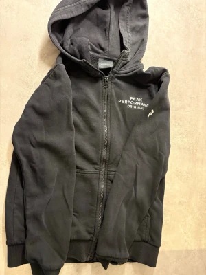 Svart hoodie från Peak Performance - Svart hoodie med dragkedja från Peak Performance. Klassisk modell med huva, fickor framtill och vit logotyp på bröstet. Perfekt för en avslappnad stil och enkel att matcha med andra plagg.