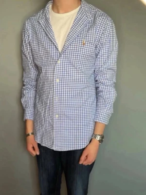 Blårutig skjorta från Ralph Lauren - Säljer en klassisk blårutig skjorta från Ralph Lauren med lång ärm och knappar framtill. Skjortan har en vit och blå rutig design och den ikoniska loggan broderad på bröstet. Perfekt till jeans eller chinos för en clean look.