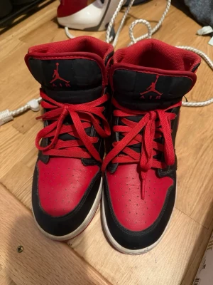 Svarta och röda Air Jordan 1 High - Säljer ett par klassiska Air Jordan 1 High sneakers i svart och rött med vita sulor. Skorna har den ikoniska Jumpman-loggan på plösen och röd swoosh på sidan. Ovandelen är i läder och mesh för en snygg och sportig look. Perfekta för dig som gillar streetwear.