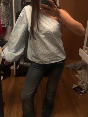 Ljusblå blommig oneshoulder-blus - Trendig ljusblå blus med blommigt mönster och oneshoulder-design. Ena ärmen är lång och puffig medan andra axeln är bar, vilket ger en cool och modern look. Finns ej att köpa längre men har Aldrig använt den!❤️ 