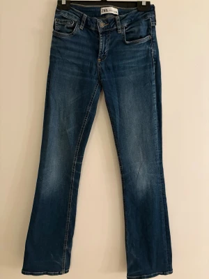 Blå bootcut jeans från ZARA - Snygga blå bootcut jeans från ZARA med klassisk femficksdesign och knappgylf. Jeansen har en lätt tvättad look och raka ben med svag utsvängning. Perfekta för en trendig och avslappnad stil.