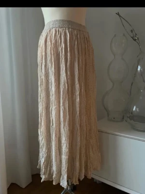Beige långkjol från NomyFicy - Lång, beige kjol från NomyFicy med elastisk midja och ett skirt, krinklat tyg. Kjolen har en lätt volangig känsla och är perfekt för dig som gillar en avslappnad men ändå stilren look.