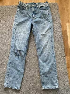 Ljusblå mom jeans med slitningar - Säljer ett par ljusblå mom jeans från HM med hög midja och raka ben. Jeansen har slitningar på vänster lår och klassiska fem fickor. Tillverkade i 100% bomull för en autentisk denimkänsla. Perfekta för en avslappnad och trendig look. Knappt använda. 