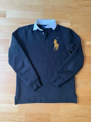 Svart långärmad skjorta från Polo Ralph Lauren - Svart långärmad skjorta från Polo Ralph Lauren med vit krage och klassisk gul broderad logga på bröstet. Skjortan har knappar vid halsen och är i en klassisk passform. Perfekt för en stilren look.