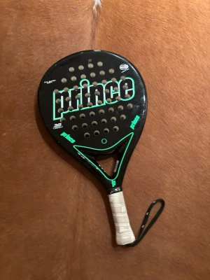 Padelracket från Prince svart/grön - Säljer ett padelracket från Prince i svart med gröna detaljer och vit lindning på handtaget. Racketen har rund form med många hål och ett handledsband. Perfekt för dig som gillar padel och vill ha en snygg och sportig look.