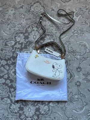 Vit Coach handväska med Snoopy - Superfin vit handväska från Coach med Snoopy och Woodstock-tryck framtill. Väskan är i läder med guldfärgade detaljer och har både axelrem och handtag. Perfekt för dig som gillar unika accessoarer och vill sticka ut med en cool design.