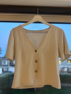 Beige kortärmad blus från 157 - Säljer en stilren beige blus från 157 med v-ringning och tre dekorativa knappar framtill. Toppen har korta ärmar och är tillverkad i ett mjukt, lätt tyg som passar perfekt till varmare dagar. Enkel och clean design som är lätt att matcha.