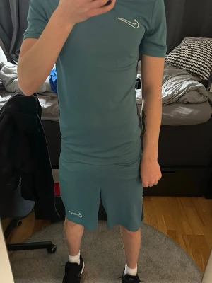Nike grön träningsset t-shirt & shorts - Säljer ett blått träningsset från Nike med matchande t-shirt och shorts. Setet har vita Nike-loggor och svarta paneler på sidorna. Perfekt för gymmet eller fotbollsträning. Materialet är lätt och andas, vilket gör det bekvämt under intensiva pass.