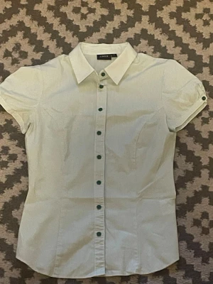 Kortärmad blus - Klassisk vit skjorta från Lindex med tunna ränder och gröna knappar. Skjortan har korta ärmar och figurnära passform, perfekt till jeans eller kjol. Snygg krage och diskret mönster ger en stilren look.
