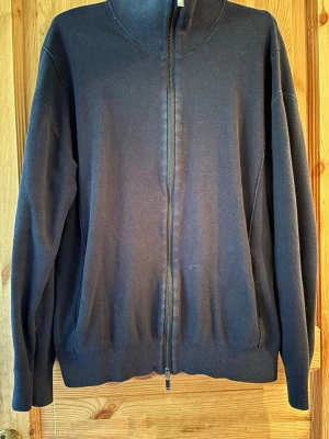 Massimo Dutti zip - En stilren svart ziptröja med hög krage och hel dragkedja framtill. Tröjan har långa ärmar, ribbade muddar och är tillverkad i ett mjukt material som passar perfekt till lager-på-lager. Enkel och clean design utan synliga loggor.