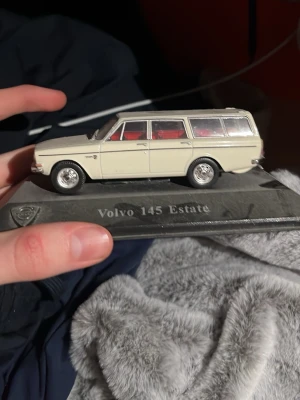 Modellbil Volvo 145 Estate vit - Säljer en detaljerad modellbil av Volvo 145 Estate i vit färg med röda säten. Bilen står på en svart platta med modellnamnet ingraverat. Perfekt för samlare eller bilentusiaster.
