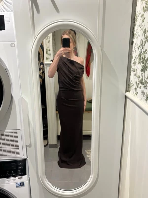 Brun långklänning oneshoulder - Snygg brun långklänning från NA-KD med oneshoulder-design. Klänningen har en elegant och figurnära passform och är tillverkad i mjuk polyester. Perfekt för dig som vill sticka ut med en stilren look.