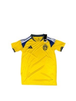 Sverige fotbollströja Adidas gul/blå - Gul fotbollströja från Adidas med blå detaljer och Sveriges landslagsemblem på bröstet. Tröjan har korta ärmar, rund halsringning med blå kant och klassiska Adidas-ränder på axlarna. Perfekt för att visa ditt stöd för Sverige på matchen!