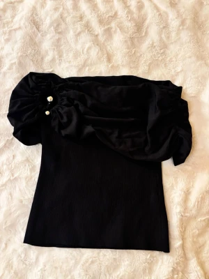 Svart offshoulder topp med pärldetalj - Trendig svart offshoulder topp med en stor puffärm och pärldetaljer på ena axeln. Toppen är ribbad och har en tight passform som framhäver figuren. Perfekt för dig som vill sticka ut med en unik och snygg topp.