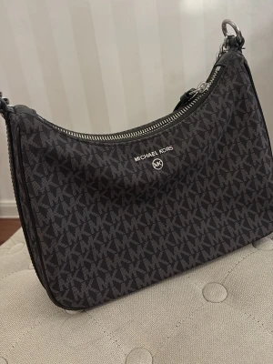 Svart Michael Kors handväska med kedja - Snygg svart handväska från Michael Kors (Äkta) med silverfärgad kedja och dragkedja. Väskan har MK-logga över hela ytan och är perfekt för att lyfta din outfit. Klassisk form och stilren design med tydlig logotyp framtill.