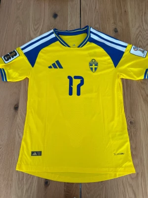 Sverige fotbollströja Gyökeres 17 - Gul och blå officiell Sverige fotbollströja från Adidas med nummer 17 och namnet Gyökeres på ryggen. Tröjan har blå detaljer, broderad landslagssköld och Climacool-material för bra ventilation. Perfekt för dig som vill stötta landslaget på match eller träning.