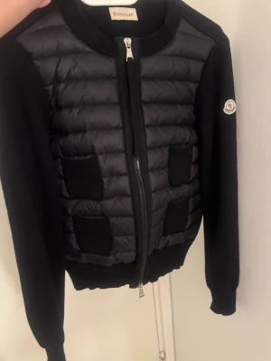 Svart cardiganjacka från Moncler - Snygg svart cardiganjacka från Moncler med quiltad framsida och två fickor. Jackan har rund halsringning, dragkedja och Moncler-logga på ärmen. Ärmar och rygg är i mjukt tyg medan framsidan är vadderad för extra stil.