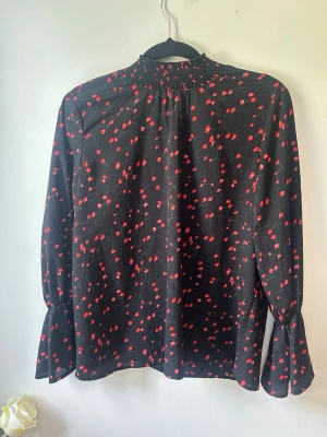 Svart blus med röda blommor - Snygg svart blus med små röda blommor och hög krage. Blusen har långa ärmar med lätt utsvängda ärmslut och ett luftigt, flowy material som ger en avslappnad men chic look. Perfekt att styla med jeans eller kjol.