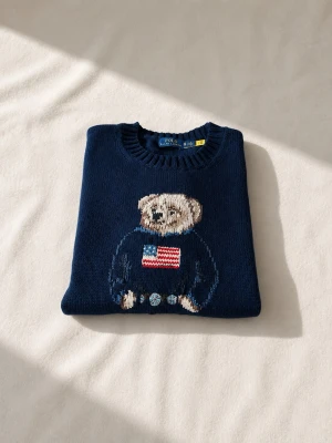 Mörkblå Polo Ralph Lauren björntröja - Mörkblå stickad tröja från Polo Ralph Lauren med ikonisk björn på bröstet, klädd i blå tröja med amerikansk flagga. Rund halsringning och ribbade muddar. RL-broderi i rött vid nederkanten. Perfekt för dig som gillar klassisk preppy-stil.