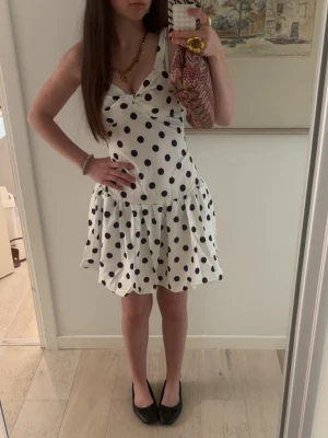 Vit prickig kort klänning - Supersöt vit klänning med svarta prickar och volangkjol. Klänningen har en axelbandslös design på ena sidan och bred axelrem på andra, vilket ger en trendig look. Perfekt för dig som gillar lekfulla och stilrena plagg.