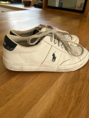 Vita Polo Ralph Lauren sneakers - Snygga vita sneakers från Polo Ralph Lauren med klassisk låg profil. Skorna har svart detalj på hälen, vita snören med svarta prickar och den ikoniska Polo-loggan på sidan. Tillverkade i läder för en clean och stilren look.