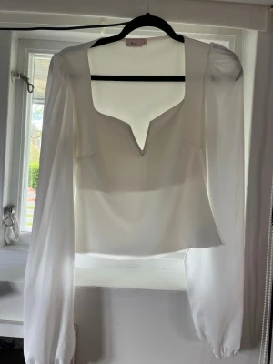 Vit blus med v-ringning från Nelly - Stilren vit blus från Nelly med djup v-ringning och långa, transparenta ärmar. Blusen har en rak passform och är tillverkad i ett lätt, flygigt material som ger en elegant look. Perfekt att matcha med både jeans och kjol.