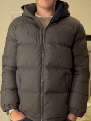 Ralph lauren jacka  - Säljer en mörkgrå dunjacka med en dragkedja framtill. Jackan har en puffig design, två sidofickor med dragkedja och elastiska muddar vid ärmsluten. Perfekt för kalla vinterdagar och riktigt skön att bära.