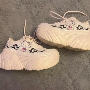 Hello Kitty platåsneakers med flames - Unika vita sneakers med extrem platåsula, Hello Kitty-tryck i rosa och svart samt svarta flames på sidorna. Skorna har snörning och coolt tryck med 'Hello Kitty' på hälen. Perfekta för dig som vill sticka ut och addera kawaii-vibe till din stil. Hely ny