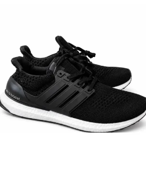 Adidas Ultra Boost svarta sneakers - Sprillans nya Svarta Adidas Ultra Boost sneakers med stickad ovandel, klassiska tre ränder på sidorna och vit Boost-sula. Skorna har snörning och en sportig, modern look. Perfekta för dig som gillar streetstyle och komfort. Materialet är mesh och syntet.
