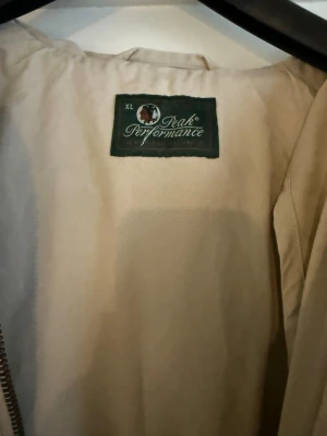 Beige vindjacka Peak Performance XL - Stilren beige vindjacka från Peak Performance med huva och dragkedja framtill. Jackan har två stora fickor med lock och är tillverkad i ett slitstarkt material som passar perfekt för blåsiga dagar. Enkel och klassisk design som funkar till det mesta.