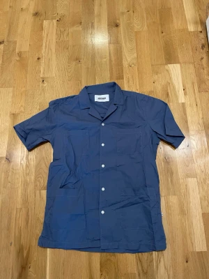 Blå kortärmad arbets skjorta från Minimum - Stilren blå skjorta från Minimum med korta ärmar och klassisk krage. Skjortan har vita knappar framtill och två bröstfickor. Perfekt för en avslappnad look och tillverkad i mjuk bomull.