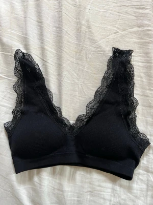 Svart spetsbralette med v-ringning - En svart bralette med djup v-ringning och dekorativa spetsdetaljer längs kanterna. Mjuk och stretchig modell utan byglar, perfekt för en bekväm och stilren look. Axelbanden är breda och täckta med spets för extra detalj.