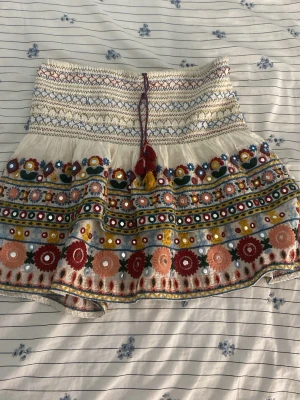 kortkjol - Superfin kortkjol i vitt med färgglada broderier och speglar. Kjolen har snörning med tofsar i midjan och volangkant. Perfekt för dig som gillar boho-stil och vill sticka ut med detaljer och mönster.