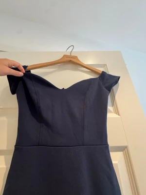 Marinblå offshoulder långklänning - Snygg marinblå långklänning med offshoulder-modell och figurnära passform. Klänningen har en v-ringad överdel och volangdetalj nertill som ger en elegant siluett. Perfekt för speciella tillfällen.
