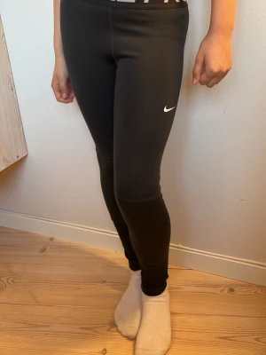 Svarta träningstights från Nike - Säljer ett par svarta träningstights från Nike med den klassiska vita swoosh-loggan på låret. Tightsen har hög midja och är tillverkade i ett stretchigt, mjukt material som passar perfekt för gym, löpning eller annan träning.
