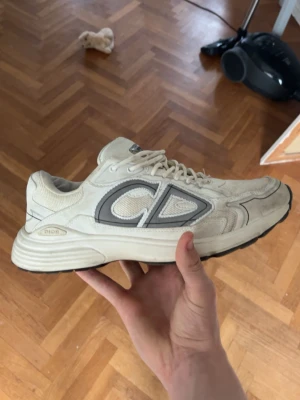 HM sneakers med CD - Säljer ett par beige HMsneakers med stora grå CD . Skorna har en chunky sula, mesh och läderpaneler samt snörning framtill. Perfekta för dig som gillar exklusiva och trendiga sneakers med streetwear-vibe.