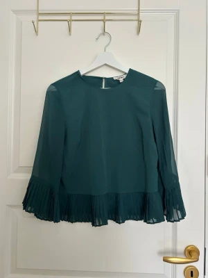 Mörkgrön plisserad blus  - Stilren mörkgrön blus från Vero Moda med transparenta långärmar och plisserade detaljer vid ärmslut och nederkant. Blusen har rund halsringning och knäppning bak i nacken. Tillverkad i ett lätt och luftigt material som ger en elegant look.