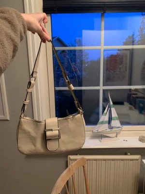 Beige axelremsväska med spänne - Stilren beige handväska i rektangulär form med justerbar axelrem och stort dekorativt spänne framtill. Väskan är tillverkad i slätt material och har minimalistisk design som passar till många outfits.