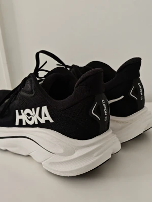 Svarta Hoka Clifton 10 sneakers 44 nya - Snygga svarta Hoka Clifton 10 sneakers med vit tjock sula och stora vita HOKA-loggor på sidan. Ovandelen är i meshmaterial för bra andningsförmåga och skorna har klassisk snörning. Perfekta för dig som gillar sportig och modern stil. Nyskick och endast provade en dag. För frågor släng ett mejl.