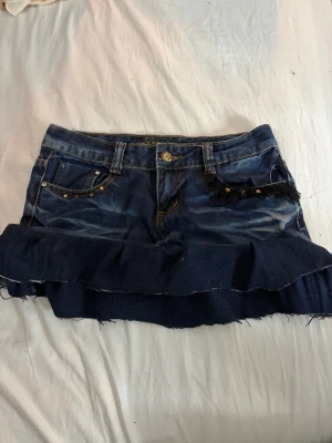 Jeanskjol med volang och nitar - Cool jeanskjol i mörkblå denim med volang nertill och snygga nitar vid fickorna. Kjolen har råa kanter och dekorativ svart spetsdetalj vid ena fickan. Perfekt för dig som gillar en edgy och trendig stil.