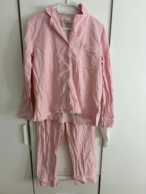 Ljusrosa pyjamas med vita detaljer - Mysig tvådelad pyjamas i ljusrosa bomull med vita passpoaldetaljer. Skjortan har krage, knappstängning och bröstficka, byxorna har raka ben. Perfekt för slappa hemmakvällar.